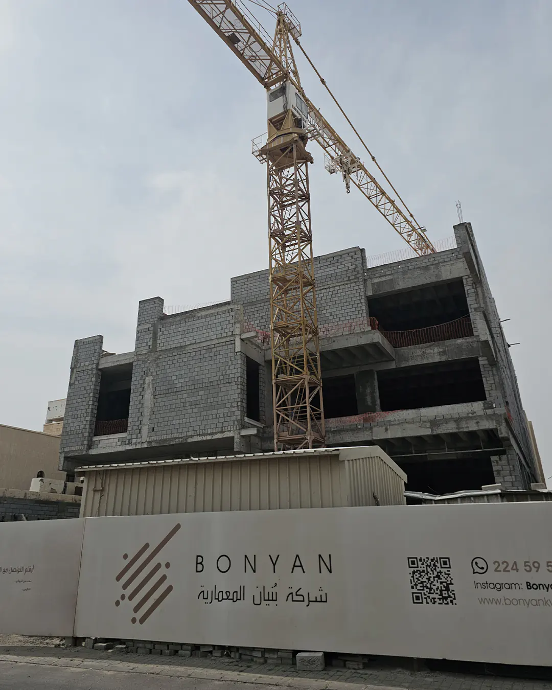 Low-angle corner view of a multi-storey unfinished villa with exposed concrete slabs, gray blockwork walls, open balcony bays, and a tower crane positioned in front of the structure behind branded site fencing.