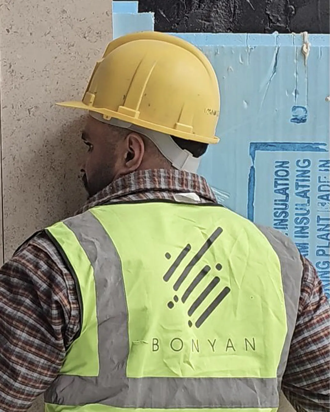 Back view of a construction worker wearing a yellow hard hat and high-visibility vest with Bonyan logo, installing exterior wall cladding panels at a job site.