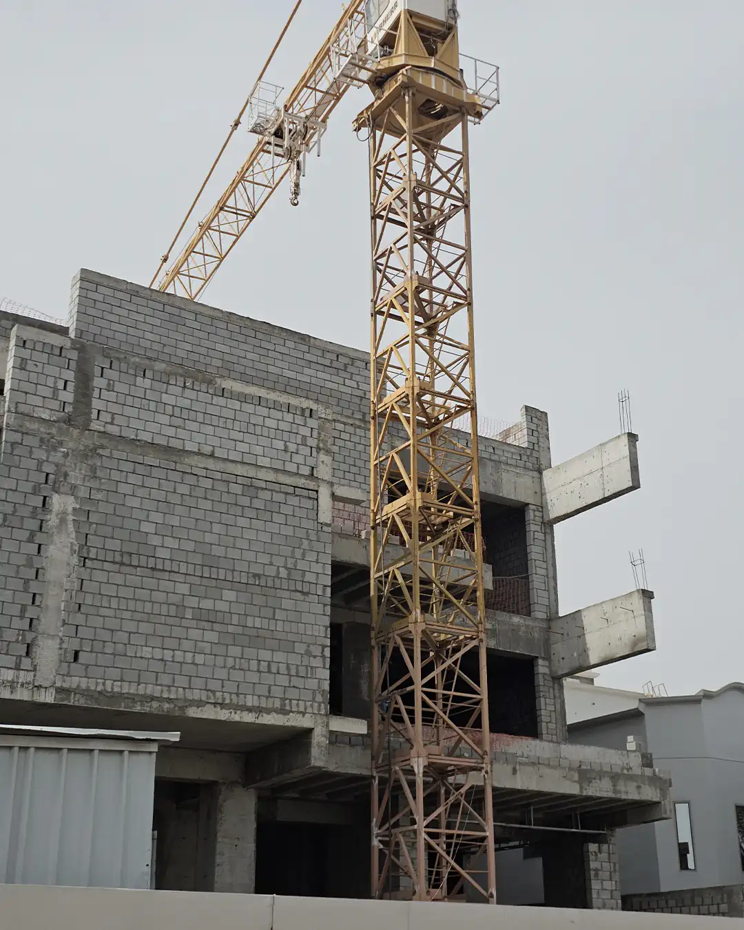 Close-up exterior view of an unfinished multi-storey villa with gray blockwork walls, exposed concrete floor slabs, projecting balcony edges, and a yellow tower crane mast standing directly in front of the structure.