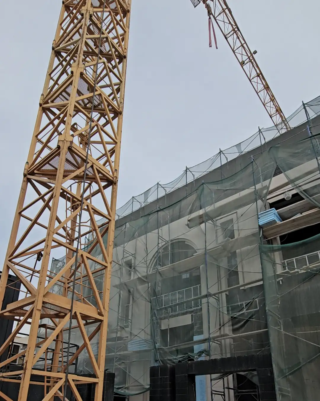 Low-angle exterior view of a villa under construction with a partially finished facade, arched upper window, scaffold mesh across the front, and a yellow tower crane and jib extending overhead.