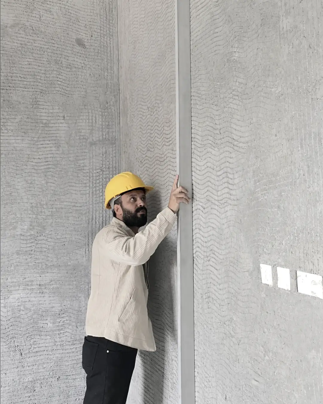 A man wearing a yellow hard hat stands inside an unfinished concrete space, checking a tall vertical frame against a textured gray wall.