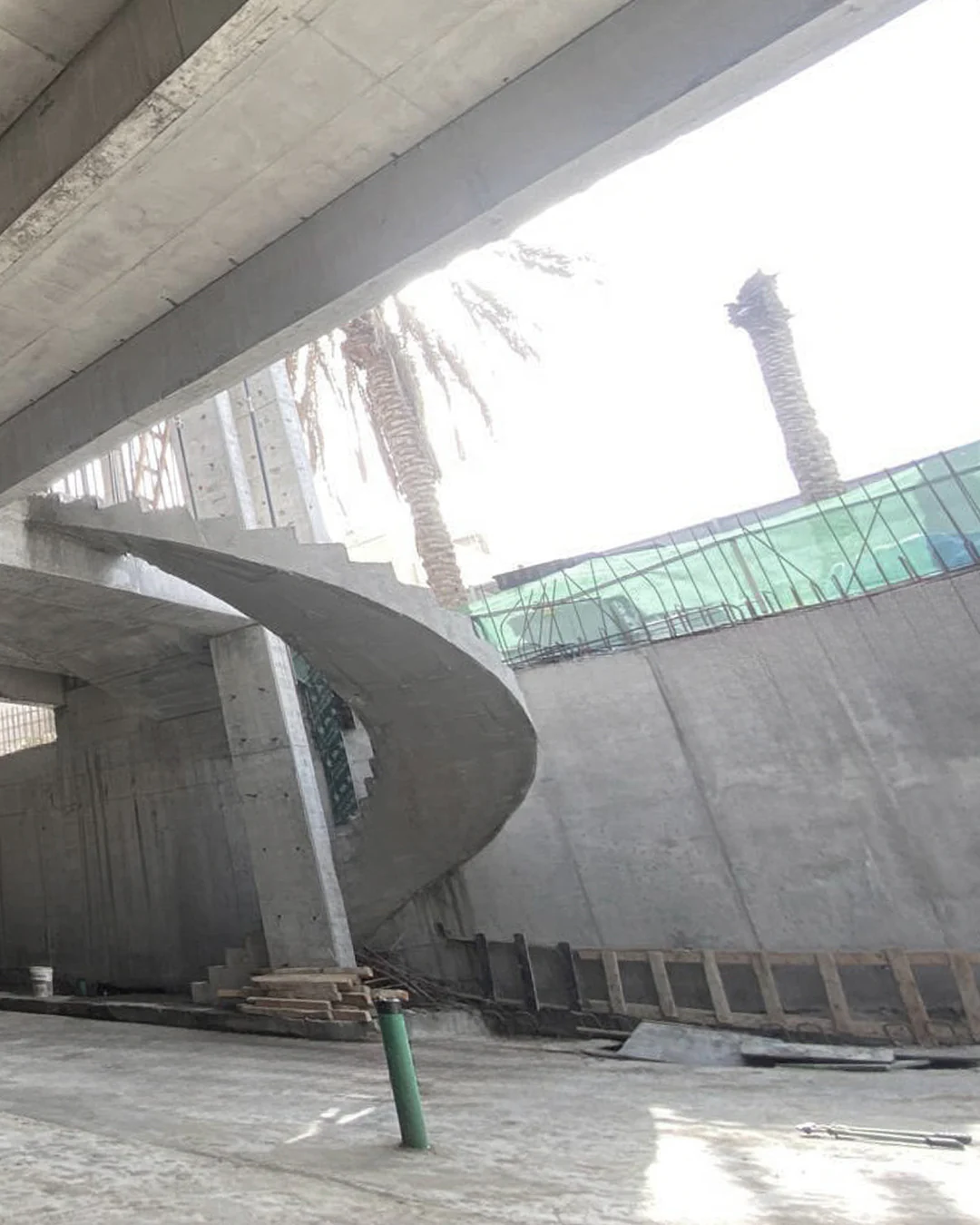Interior view of a reinforced concrete building featuring a curved staircase, structural columns, slab beams, and surrounding foundation walls under construction.