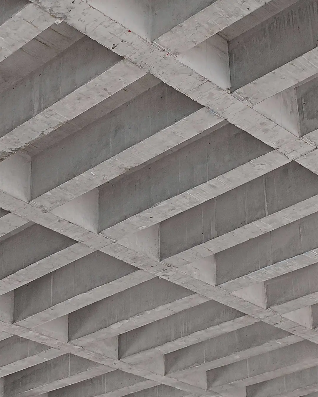 Close-up view of an unfinished reinforced concrete ceiling with intersecting beams forming a square grid pattern.