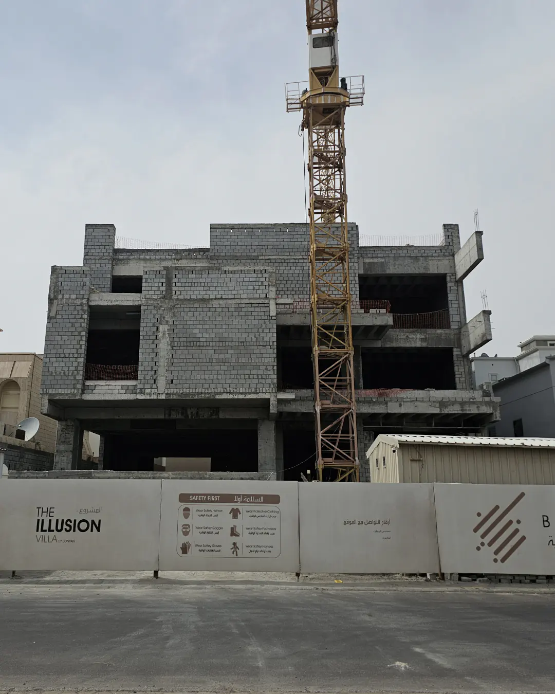 Street-facing view of a multi-storey unfinished concrete villa with blockwork walls, open floor bays, a tower crane rising through the center, and site fencing branded “The Illusion Villa” along the front.