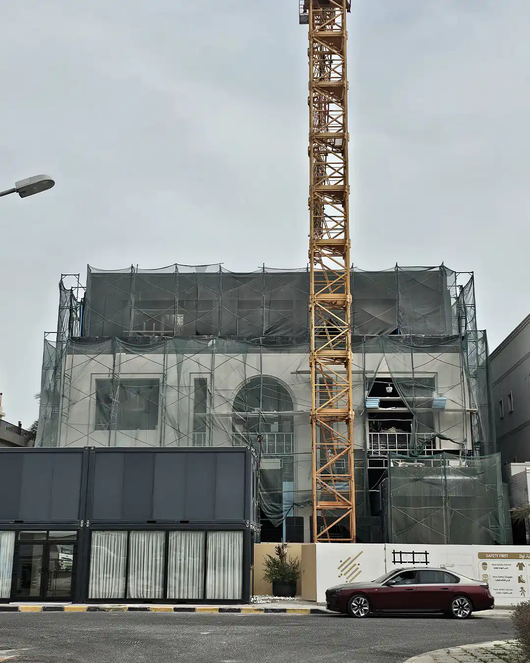 Street-facing view of a multi-storey villa under construction with a partially finished white facade, scaffold mesh wrapping the upper levels, a tower crane in front, and site fencing along the road.
