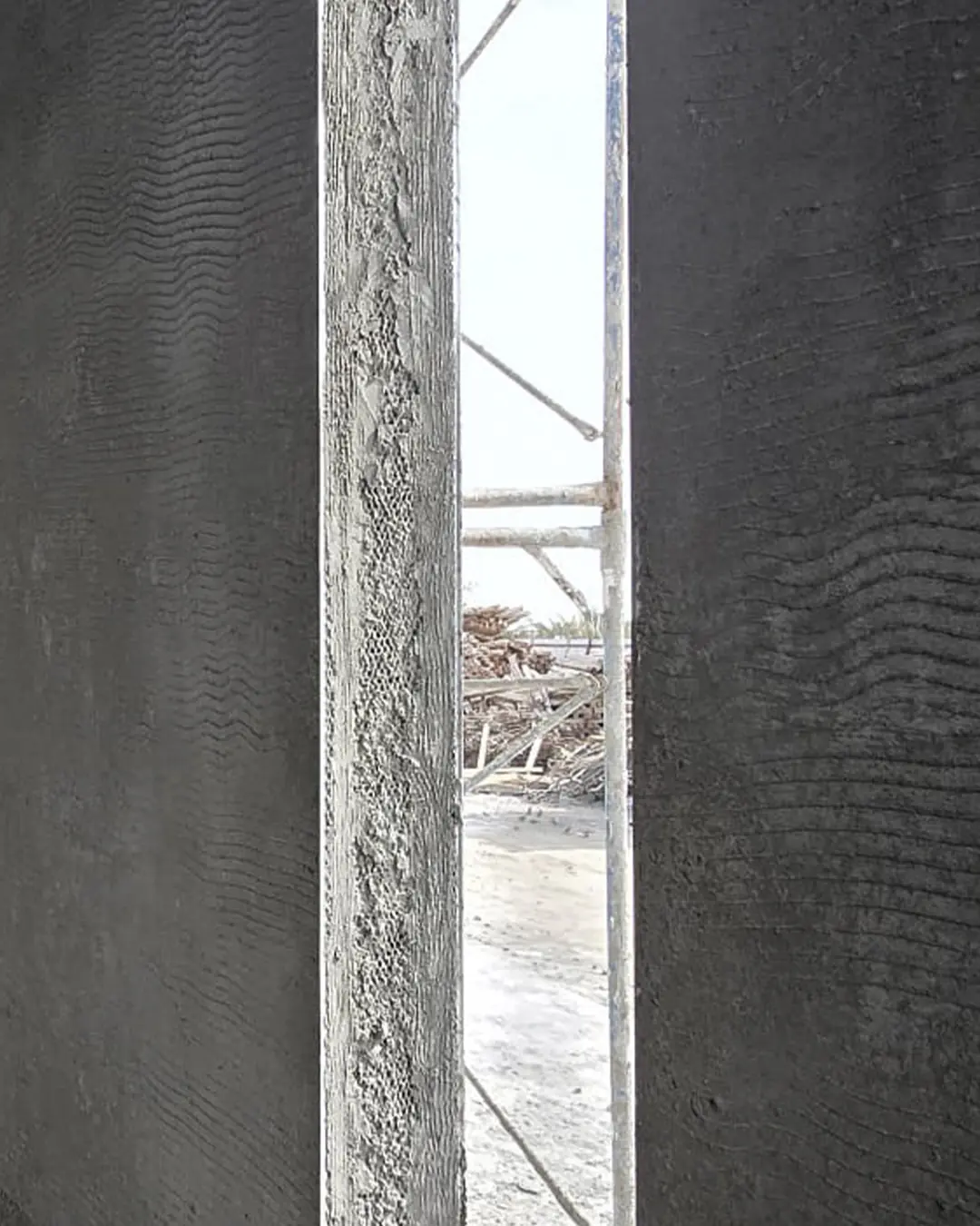 Close-up view through a slim vertical opening between dark textured concrete walls, revealing scaffolding and construction debris outside in bright daylight.