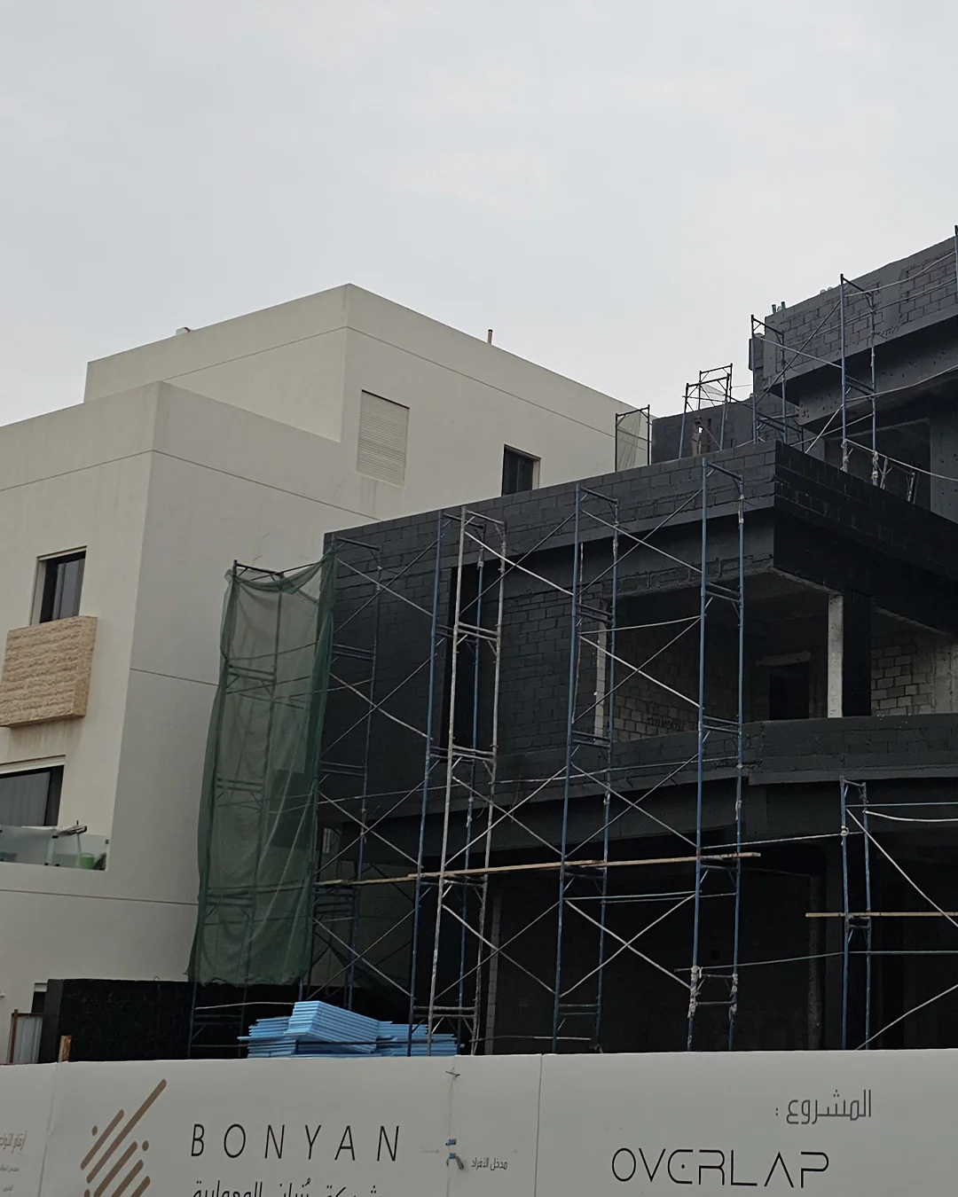Side view of a modern two-story villa under construction with exposed concrete blocks and metal scaffolding, adjacent to a finished white building, branded fencing reading “Bonyan” and “Overlap.”