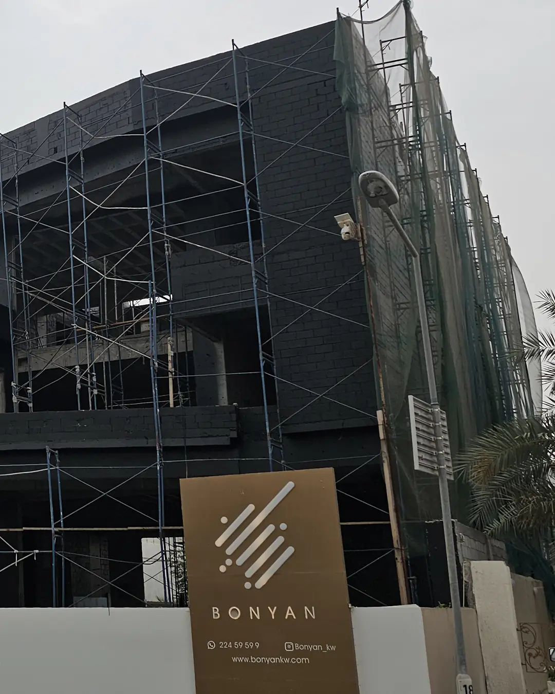 Corner view of a modern two-story villa under construction with dark concrete block walls and extensive metal scaffolding, Bonyan branding sign in front, and protective green netting along the side facade.