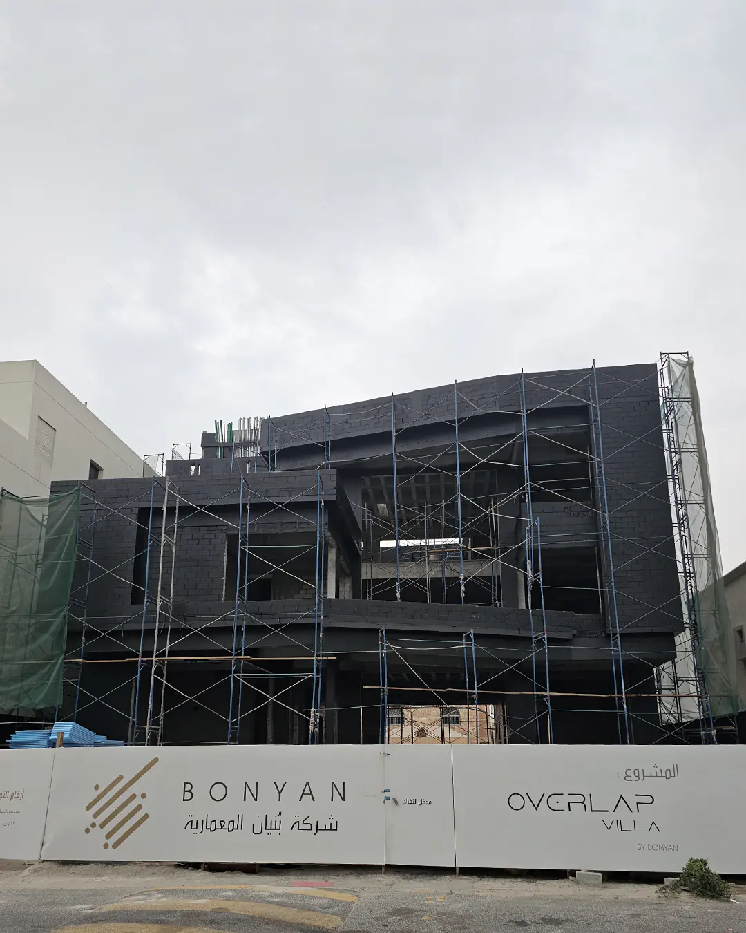 Modern two-story villa under construction with scaffolding covering the facade, branded construction fencing reading “Bonyan” and “Overlap Villa,” on a cloudy day.