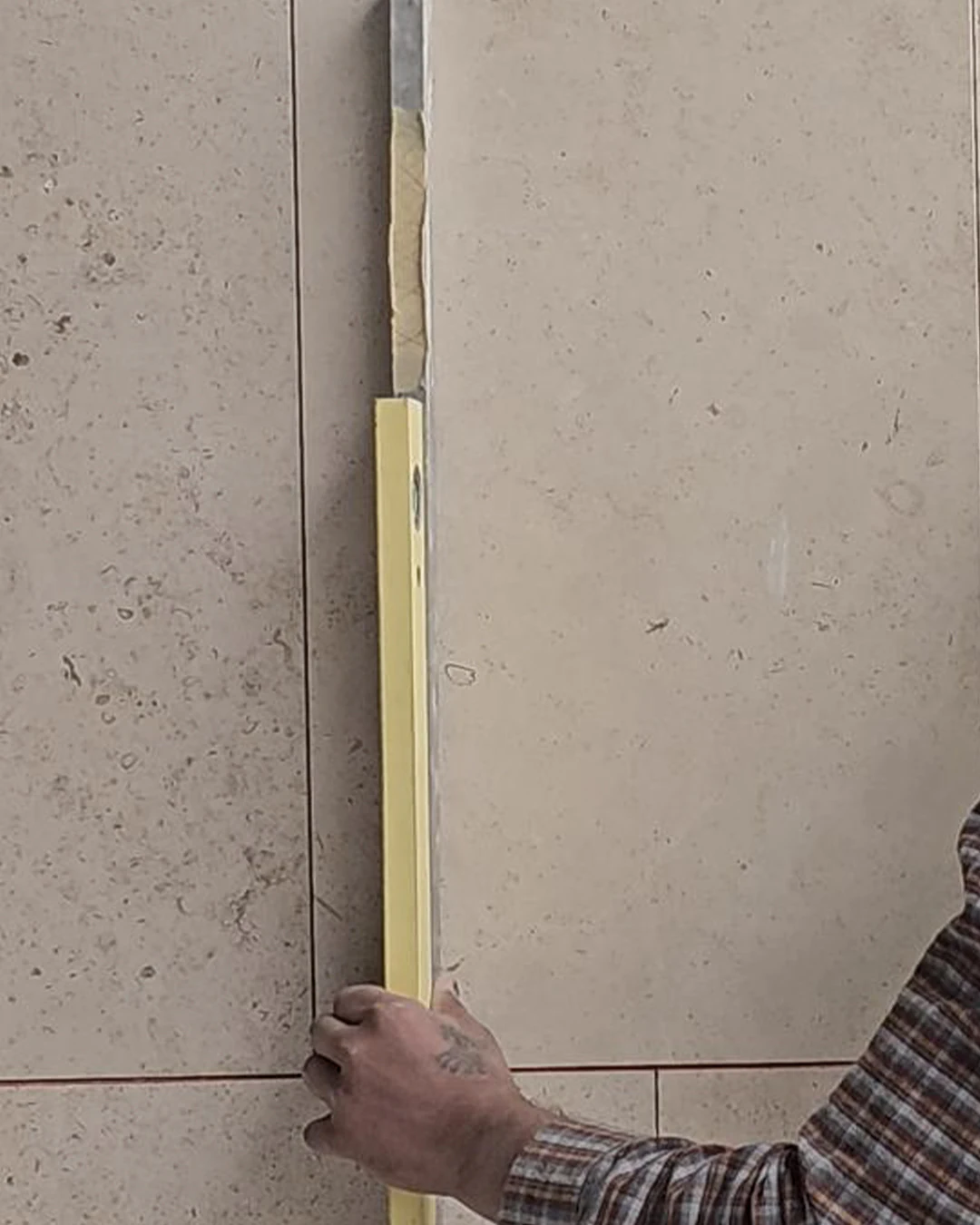 Close-up of a worker’s hand holding a spirit level vertically against a stone tile wall to verify straight alignment and even joint spacing.