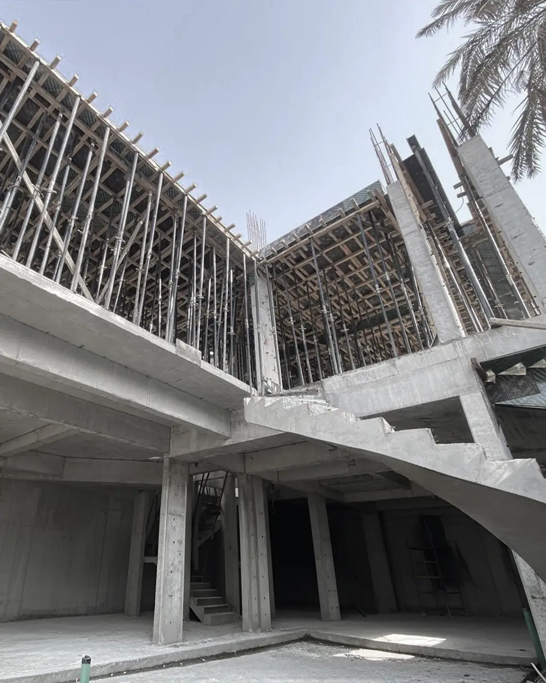 Reinforced concrete building frame showing columns, beams, a staircase, and upper-level slab formwork supported by dense scaffolding under an open sky.