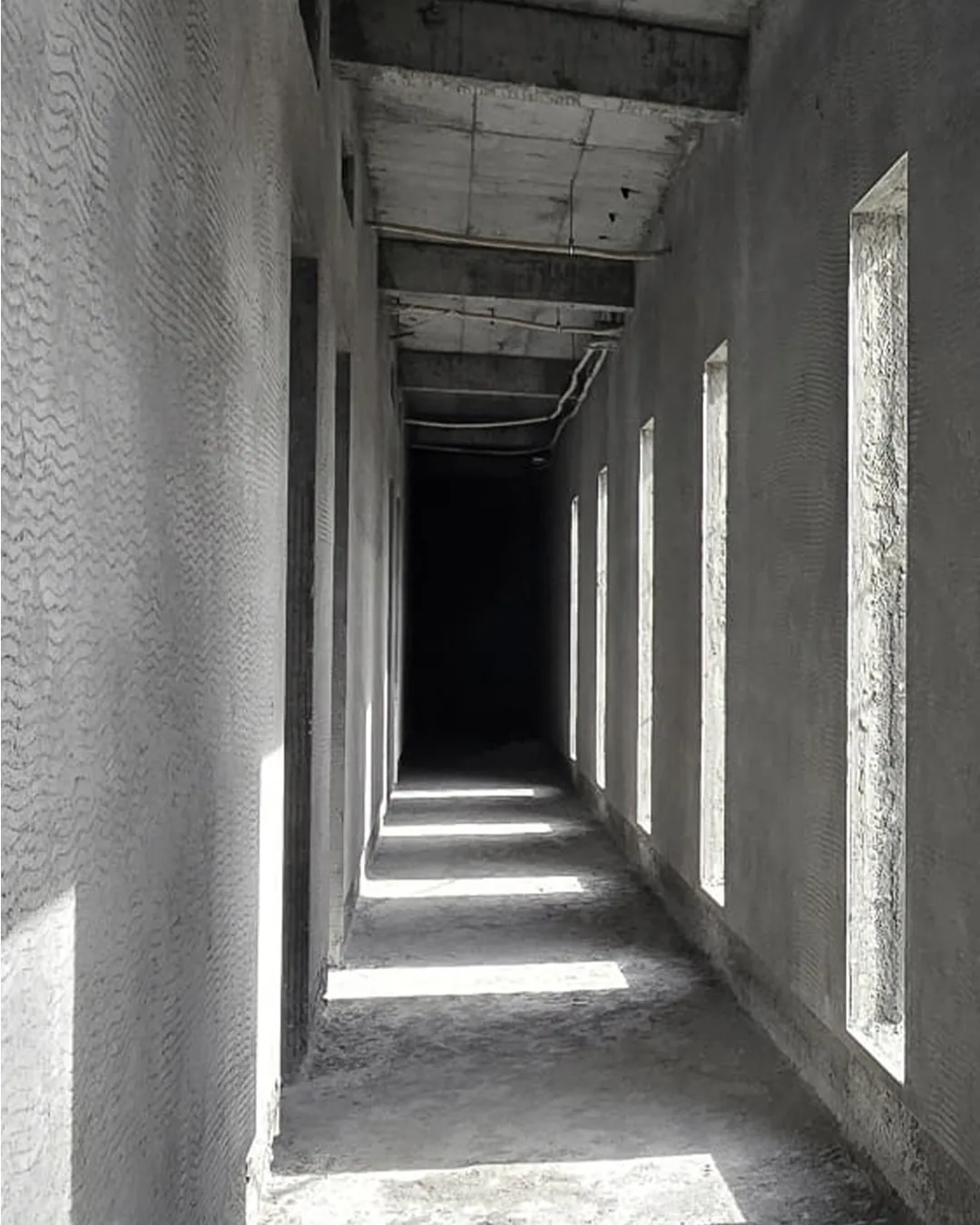 Narrow unfinished hallway with textured concrete walls, exposed ceiling beams, and tall slit windows casting bright bands of light across the floor.