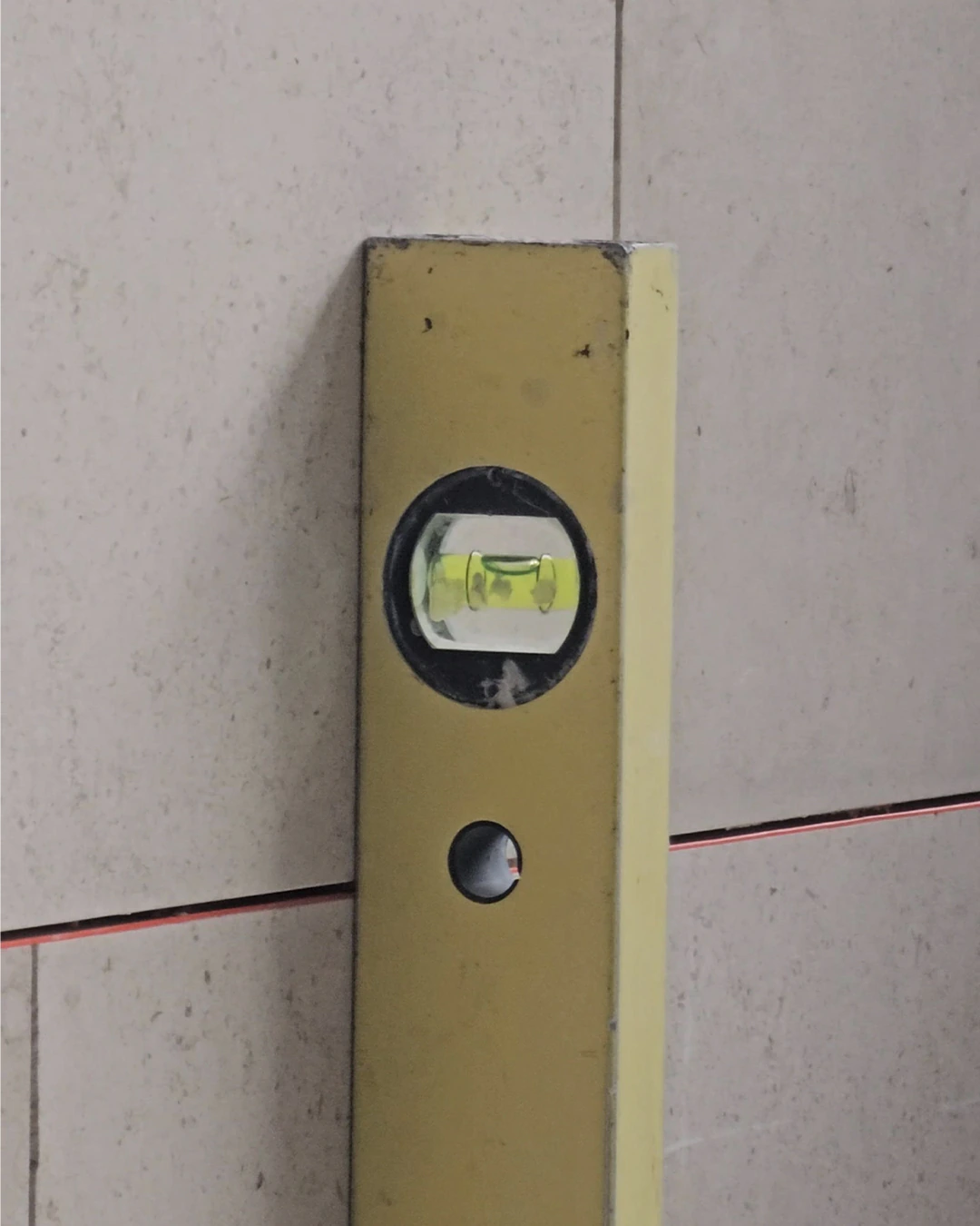 Close-up of a yellow spirit level placed vertically against a light stone tile wall, showing the bubble centered to confirm proper alignment.