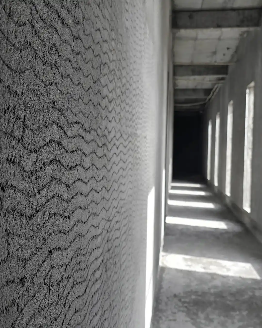 Close-up of a rough patterned concrete wall beside a narrow unfinished corridor, with tall window openings casting bright strips of sunlight onto the floor.