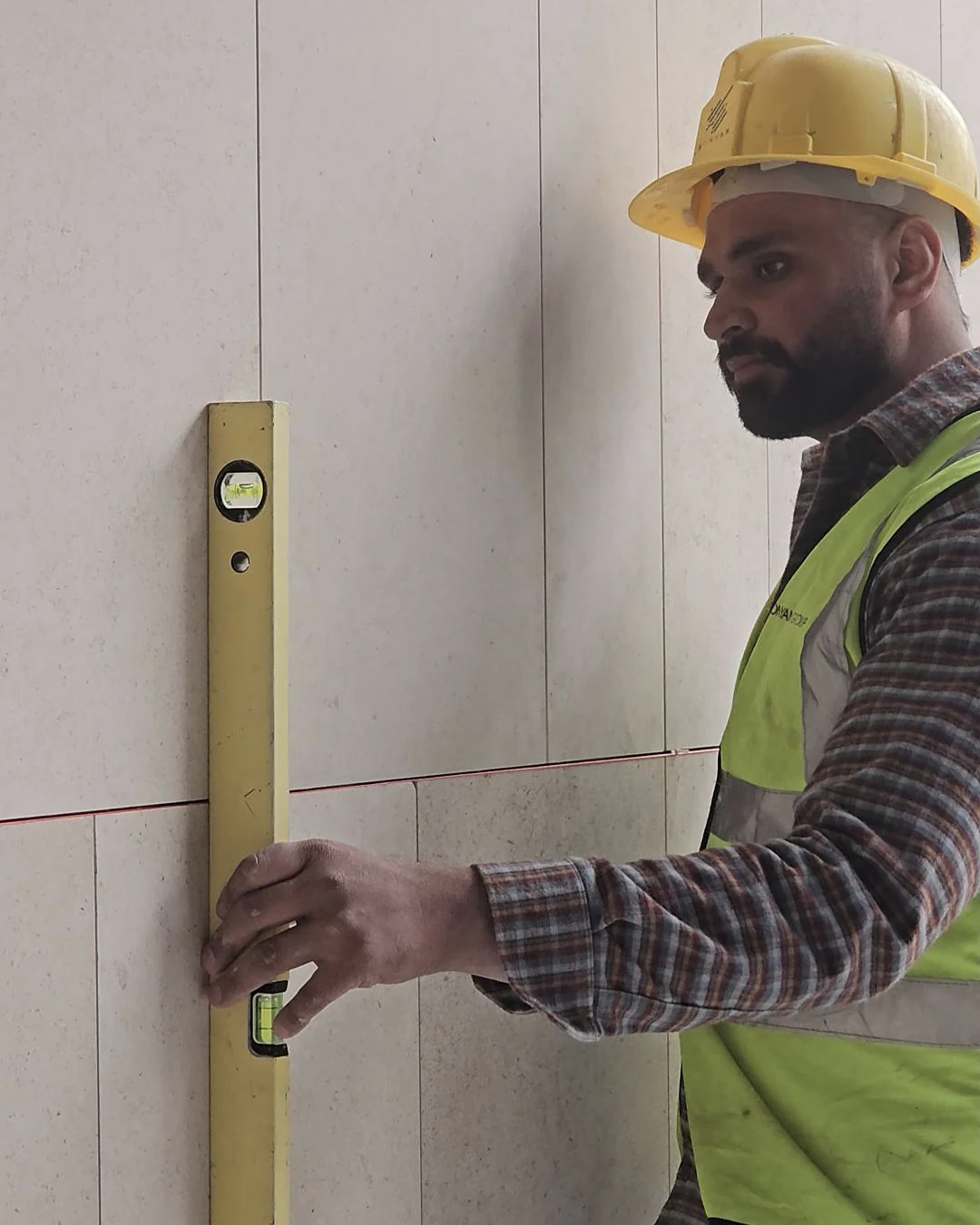 Construction worker wearing a yellow hard hat and safety vest using a spirit level to check vertical alignment on a tiled wall surface.